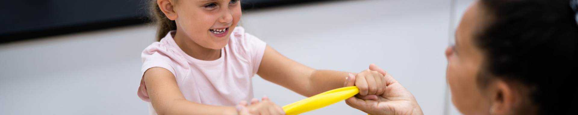 Eine Therapeutin mit einem kleinen Mädchen - gemeinsam ziehen sie ein gelbes Terraband auseinander